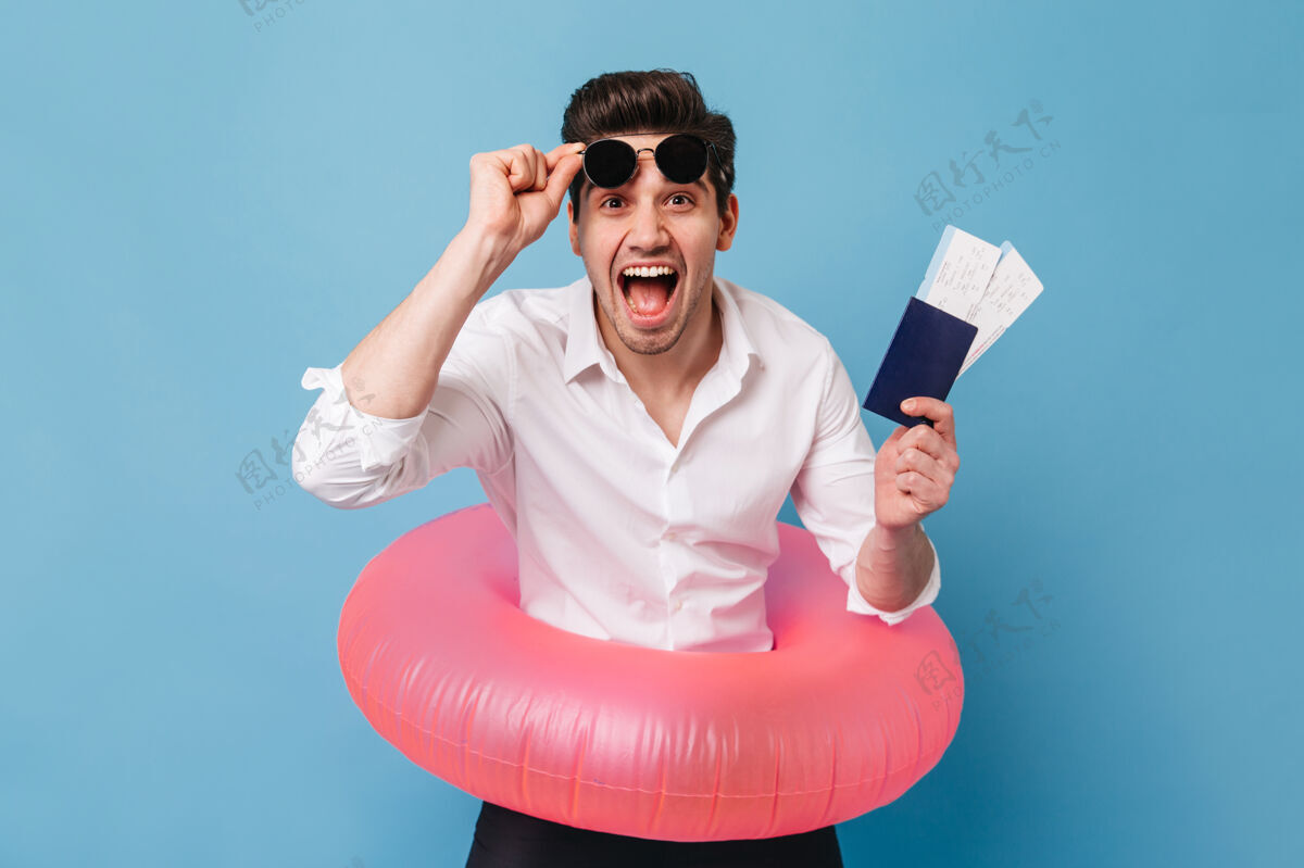 现代情绪激动的棕色眼睛的家伙摘下太阳镜 高兴地挥舞着他的护照和机票一个穿着白衬衫的男人拿着橡胶圈在蓝色空间里摆姿势男性搞笑成功