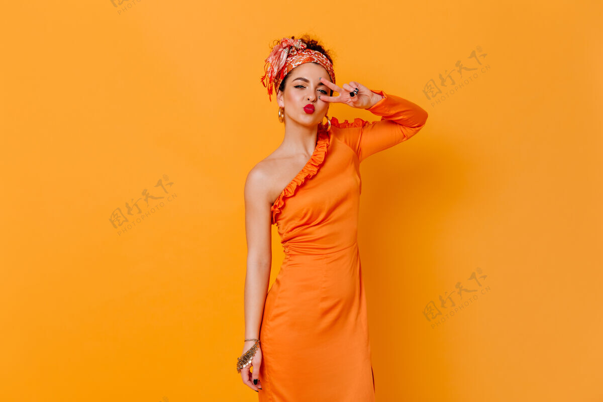 魅力身着丝绸长裙 头箍的风骚女子在橙色的空间吹吻并显示和平标志头带红色唇膏头发