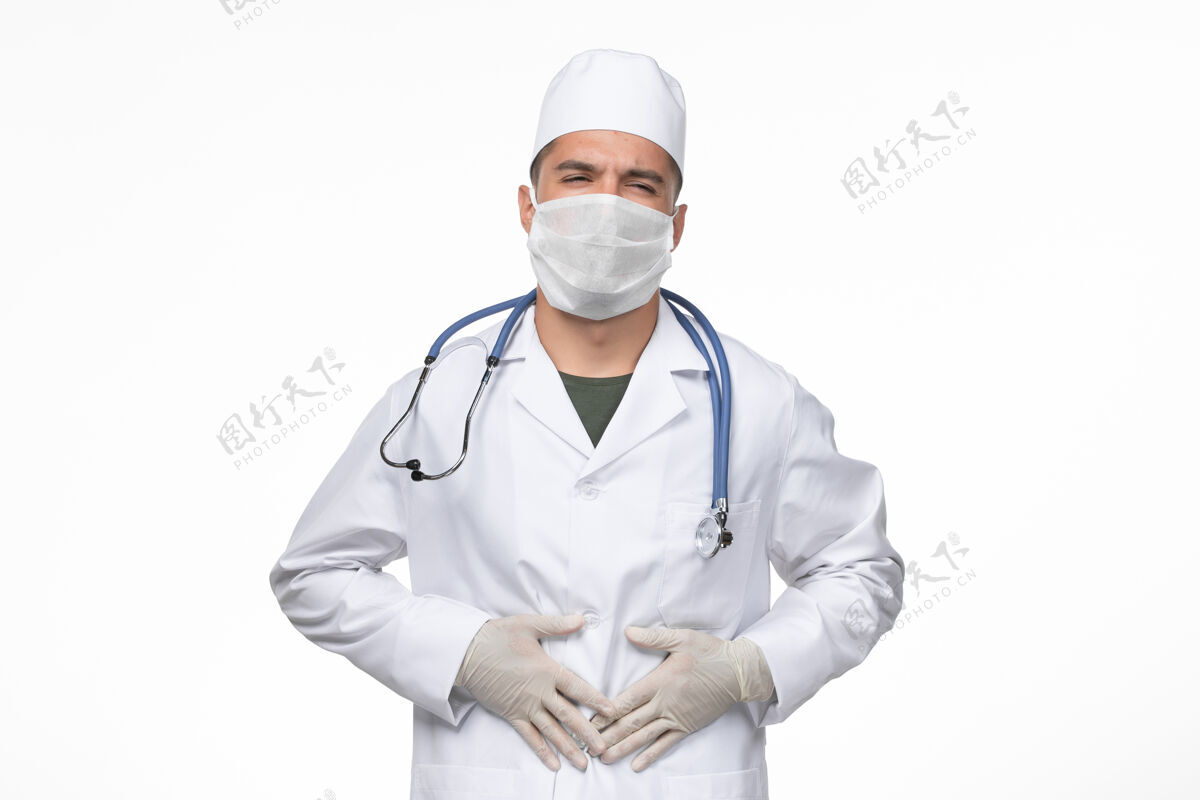 正面正面图男医生穿着医疗服 戴着口罩 在白墙上抵御胃痛冠状病毒-冠状病毒病隔离大流行观点医生面具