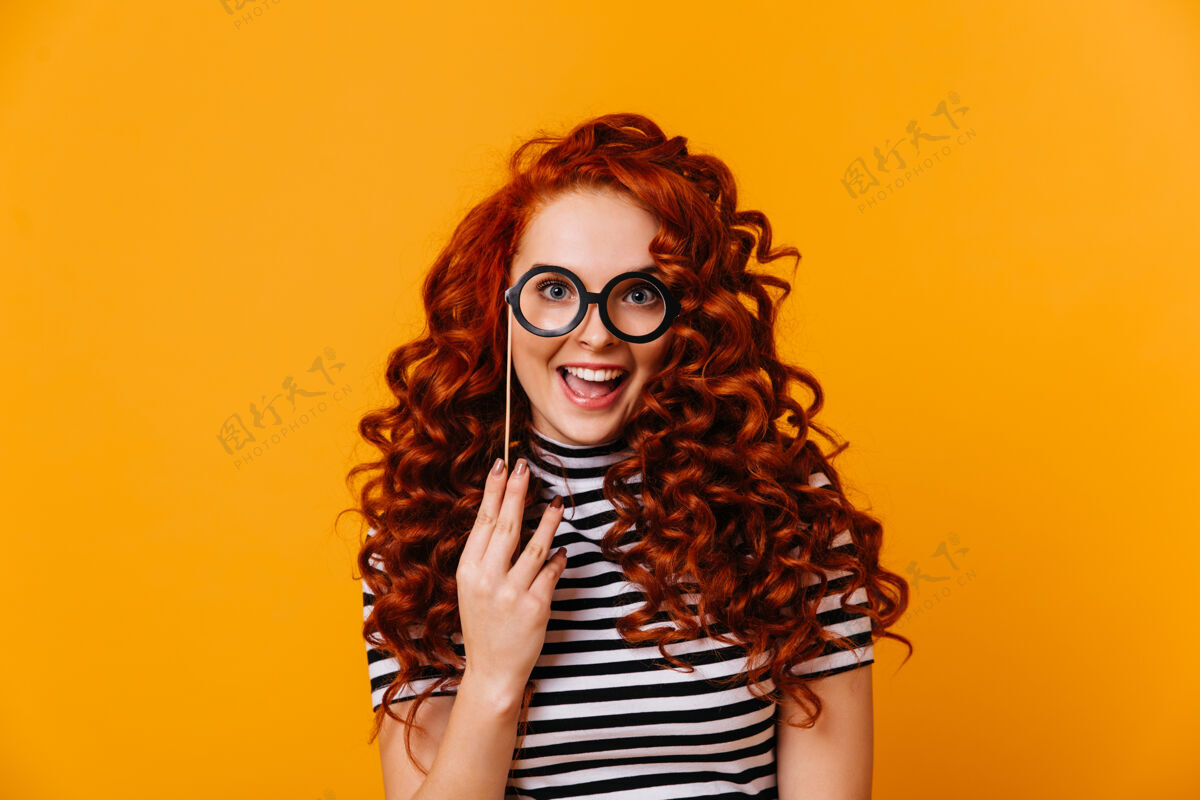 正面蓝眼睛红头发的迷人女人手持眼镜模特 在橙色的空间里微笑着条纹时尚表情