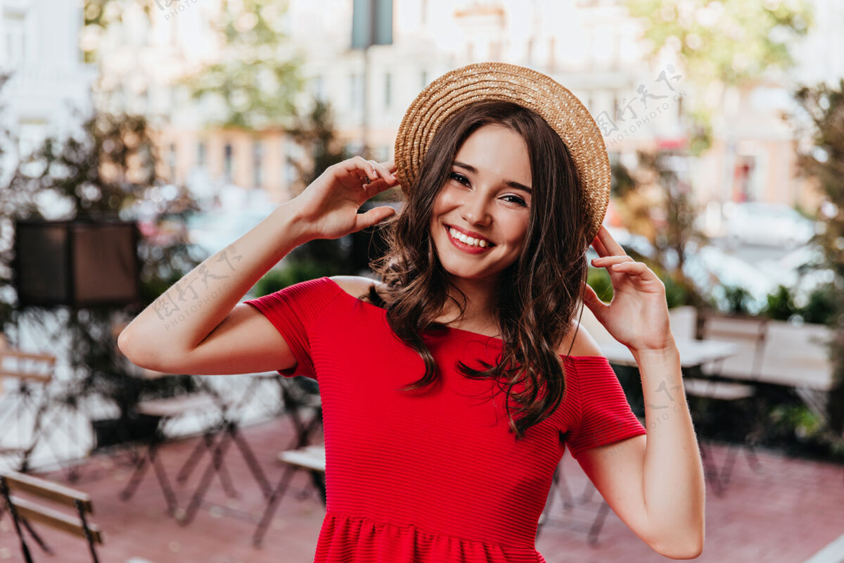 户外棕色头发的女模特戴着草帽摆姿势穿着红色连衣裙的幽默女孩站在城市的户外照片户外女人快乐