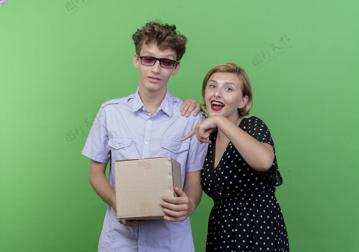 站着年轻漂亮的夫妇困惑的男子拿着盒子包 而他的女友用手指着盒子开心的微笑站在绿色的墙上情侣微笑女朋友