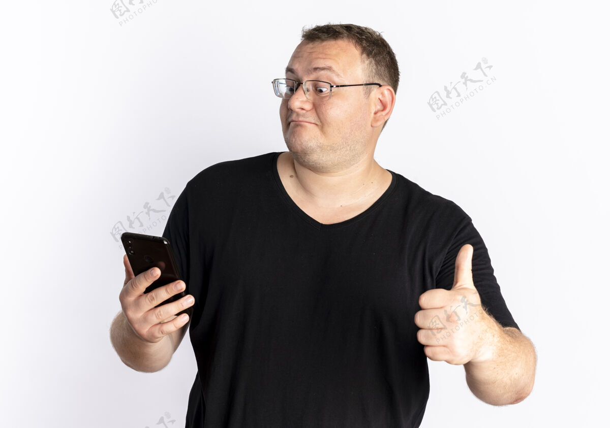 看一个戴眼镜 身穿黑色t恤的超重男子站在白色的墙上 看着他的智能手机屏幕 竖起大拇指显示手机拇指