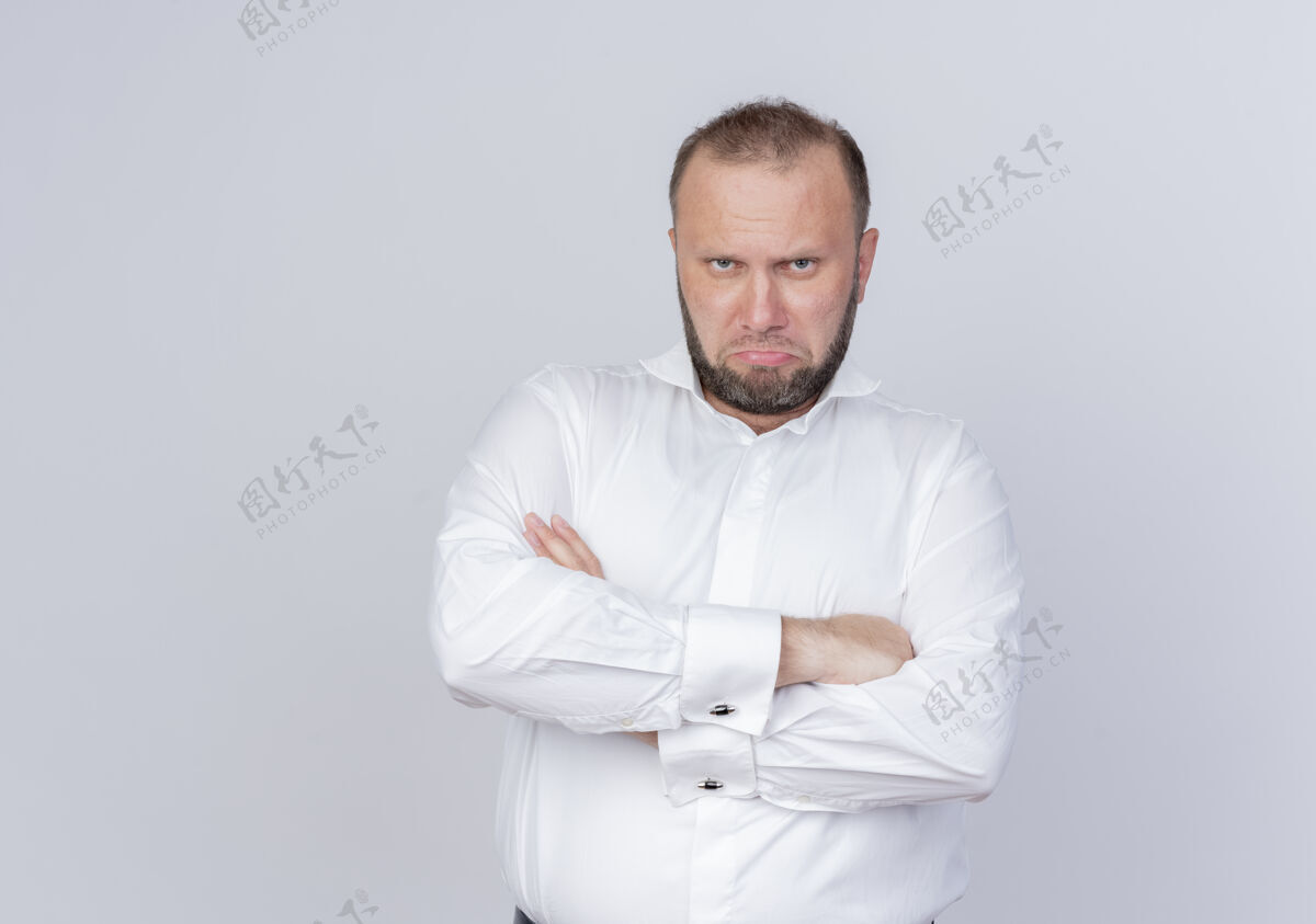 怨恨愤怒的胡子男人穿着白衬衫 严肃的脸交叉双臂站在白墙上胳膊严肃看