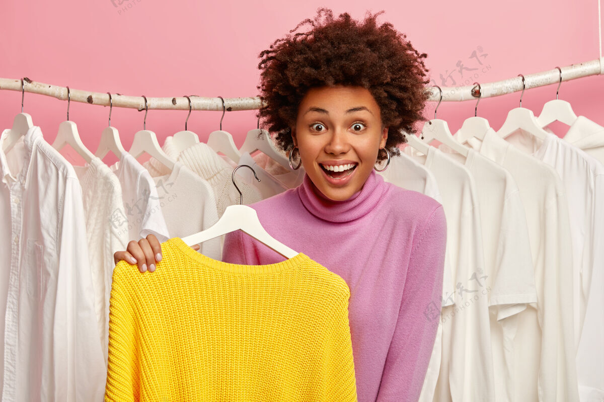 选择有着非洲发型的快乐女人 衣架上挂着亮黄色的毛衣 在打折时拿起衣服 站在衣柜旁边女士请消费主义