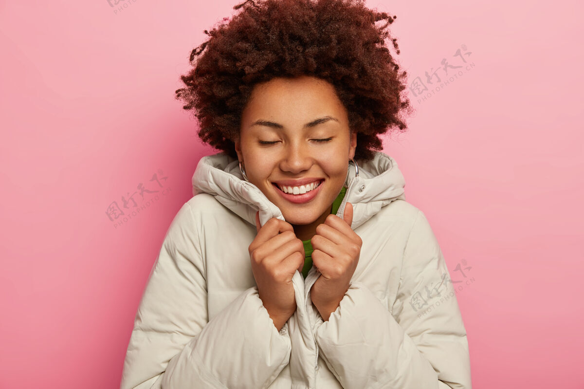 卷发快乐迷人的女孩穿着白色夹克感到温暖 笑容灿烂 眼睛紧闭 露出完美的牙齿 站在粉色的工作室背景上温暖粉色深色