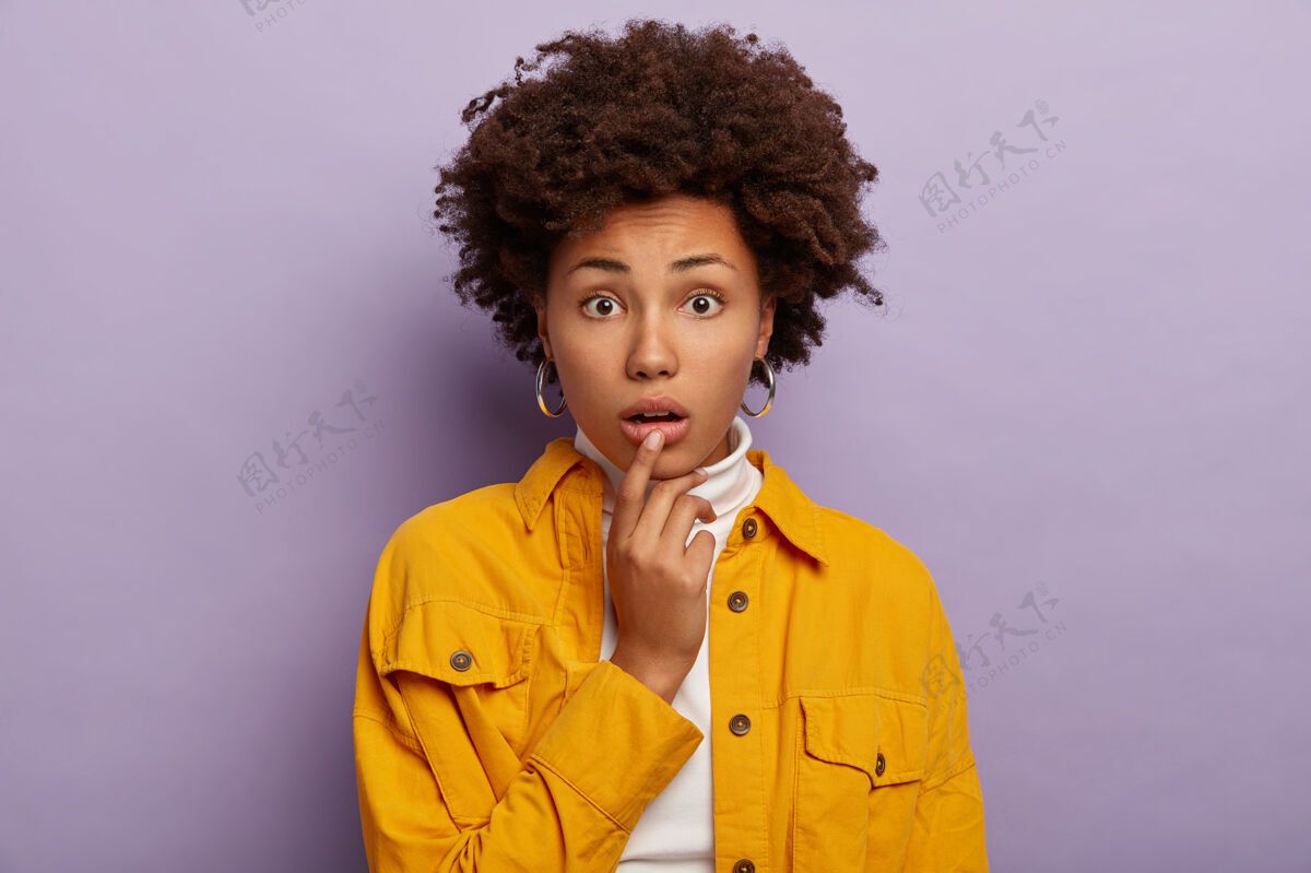 惊人神经兮兮的迷惑不解少女的画像摸着下唇 有着卷曲的非洲发型 焦虑的表情 穿着时髦的黄色夹克和耳环 模特们在紫色的背景下室内表演高领毛衣拼图皱眉