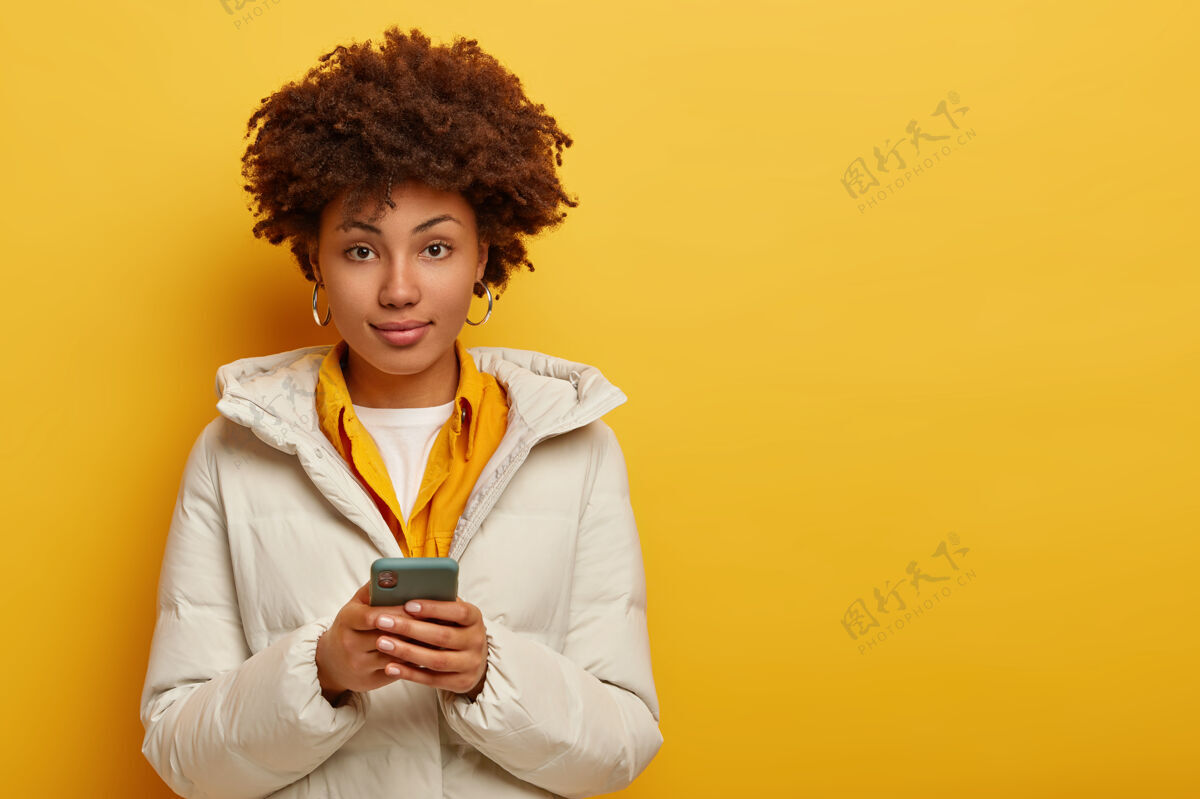 聊天穿着白色保暖外套的时尚迷人的女人 直视镜头 用现代手机上网聊天 卷发 隔离在黄色背景下人与现代科技白手机美国