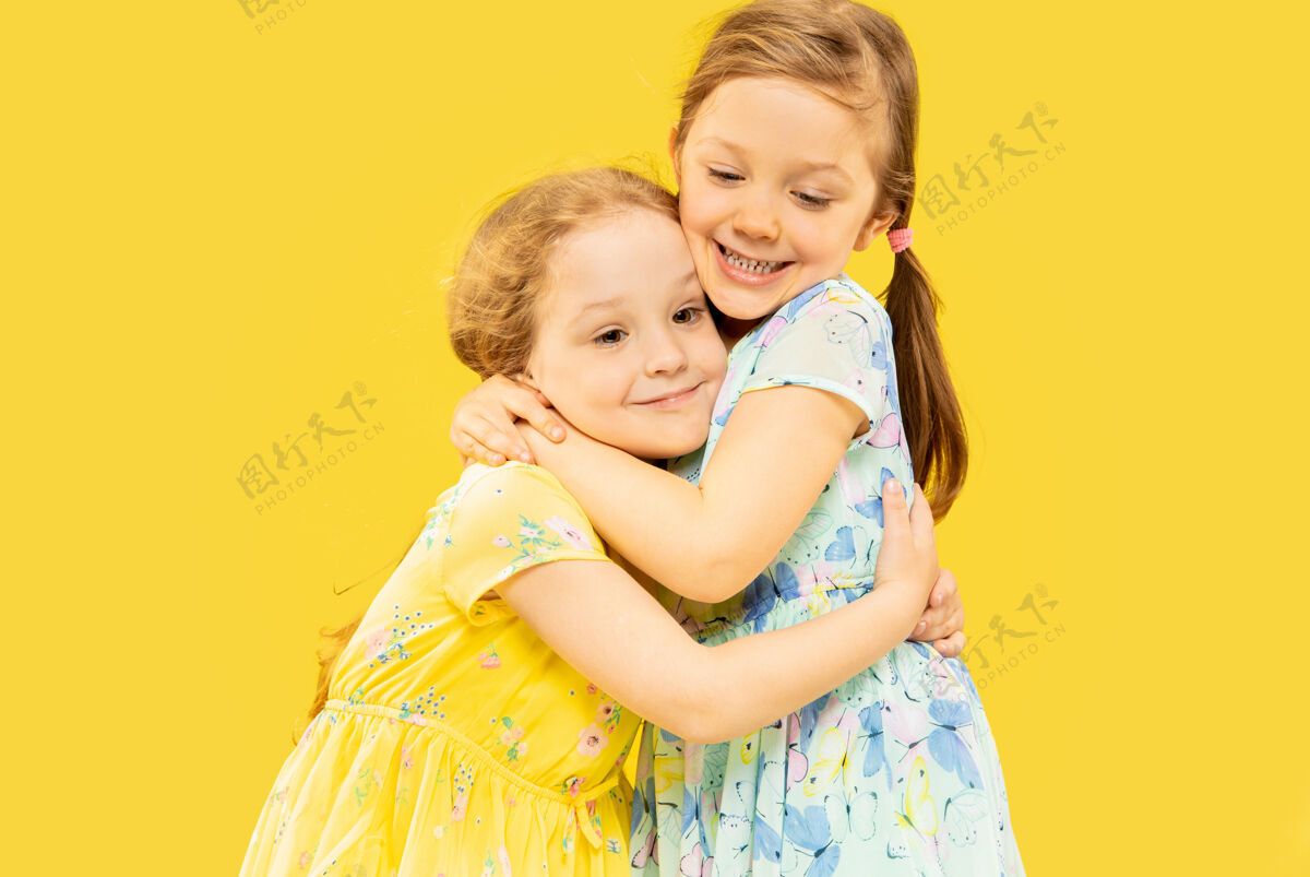 手势美丽动情的小女孩被隔离在黄色的空间里两个快乐的姐妹穿着裙子互相拥抱的半身肖像面部人类女性