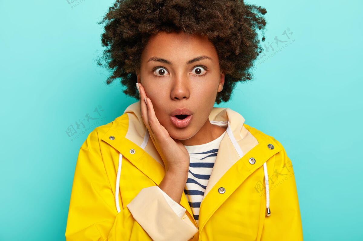黄色惊呆了的女人的照片屏住呼吸 用窃听的眼睛盯着 对令人震惊的缓和反应 有非洲发型 穿着黄色雨衣 隔离在蓝色背景下 无语休闲卷发脸颊