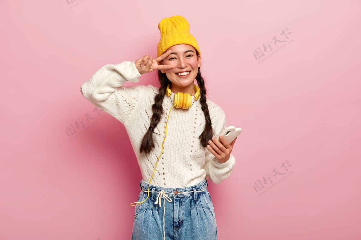 音乐笑容满面的韩国年轻女子在眼睛上做了一个和平的手势 手持现代手机 有两个辫子 微笑温柔 戴着黄色帽子和牛仔裤 在粉色背景下摆姿势牛仔裤白色室内