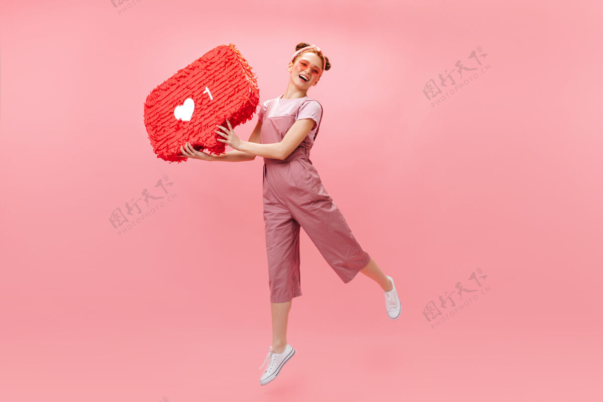 时尚快乐的女人穿着时髦的连身衣 在粉色背景上跳跃 举着像标牌一样的东西红发肖像化妆品