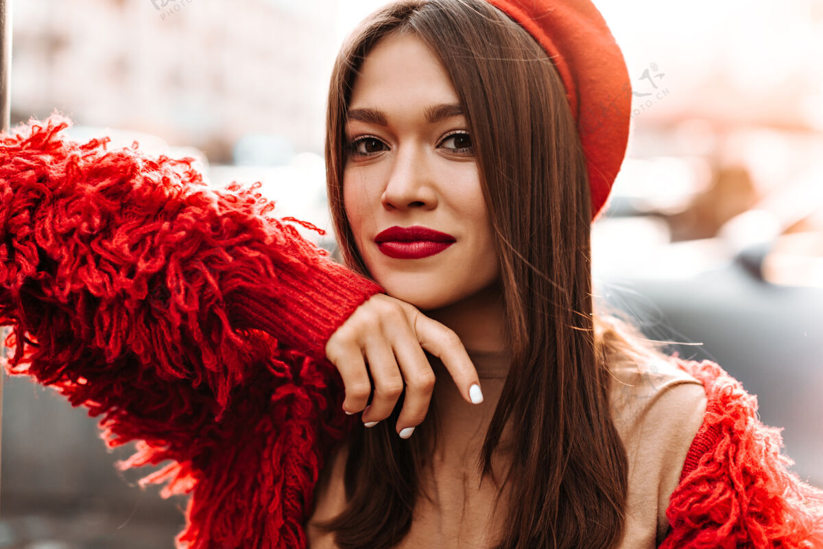 时尚一个白指甲红唇的女人在街上的背景下看着摄像机一个穿着红色贝雷帽和羊毛夹克的女人靠在窗户上阳光漂亮外面