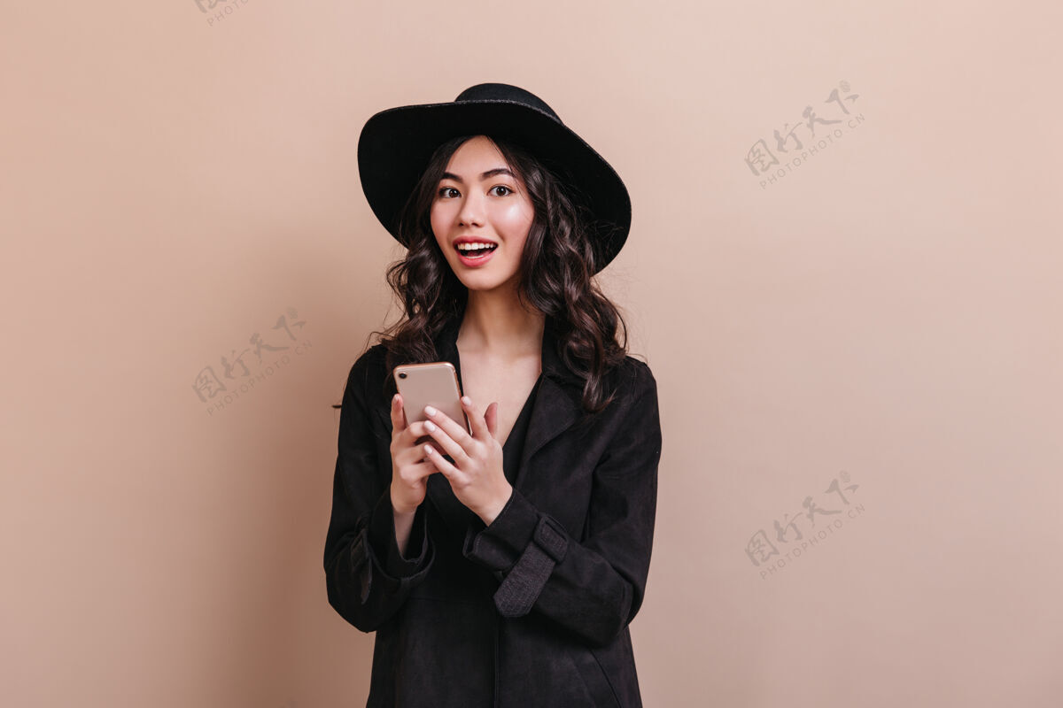 女人惊艳的亚洲女人拿着智能手机看着相机优雅的卷发女人穿着外套摆着小玩意儿中国人表情时尚