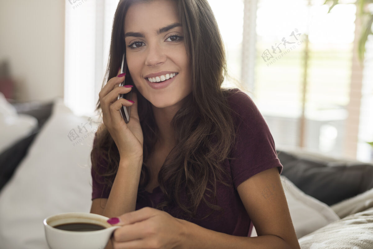 家庭内部快乐美丽的年轻女子拿着咖啡杯和电话交谈沙发忙着电话