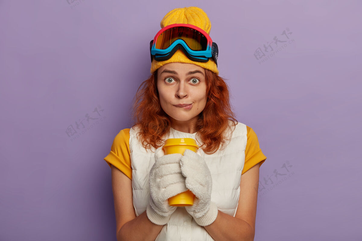 滑雪板照片中的美女模特长着姜黄色的头发 咬着嘴唇 双手捧着外卖杯 直视镜头穿着黄色t恤搭配背心手套帽子红发
