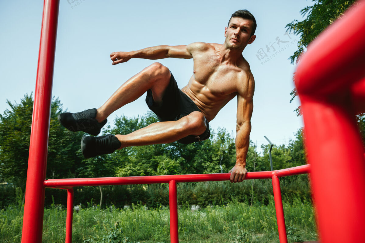 肌肉在阳光明媚的夏日 一个年轻肌肉发达 赤膊上阵的白人男子在操场上跳过单杠锻炼水平区域
