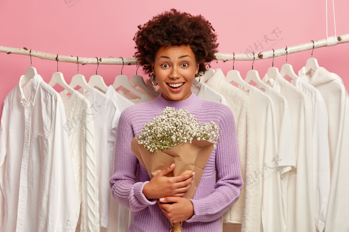 请快乐的黑皮肤女人带着花束在更衣室里摆姿势 穿着紫色毛衣 背景是衣架上的白色衣服 微笑着看着镜头紫色美国夹克