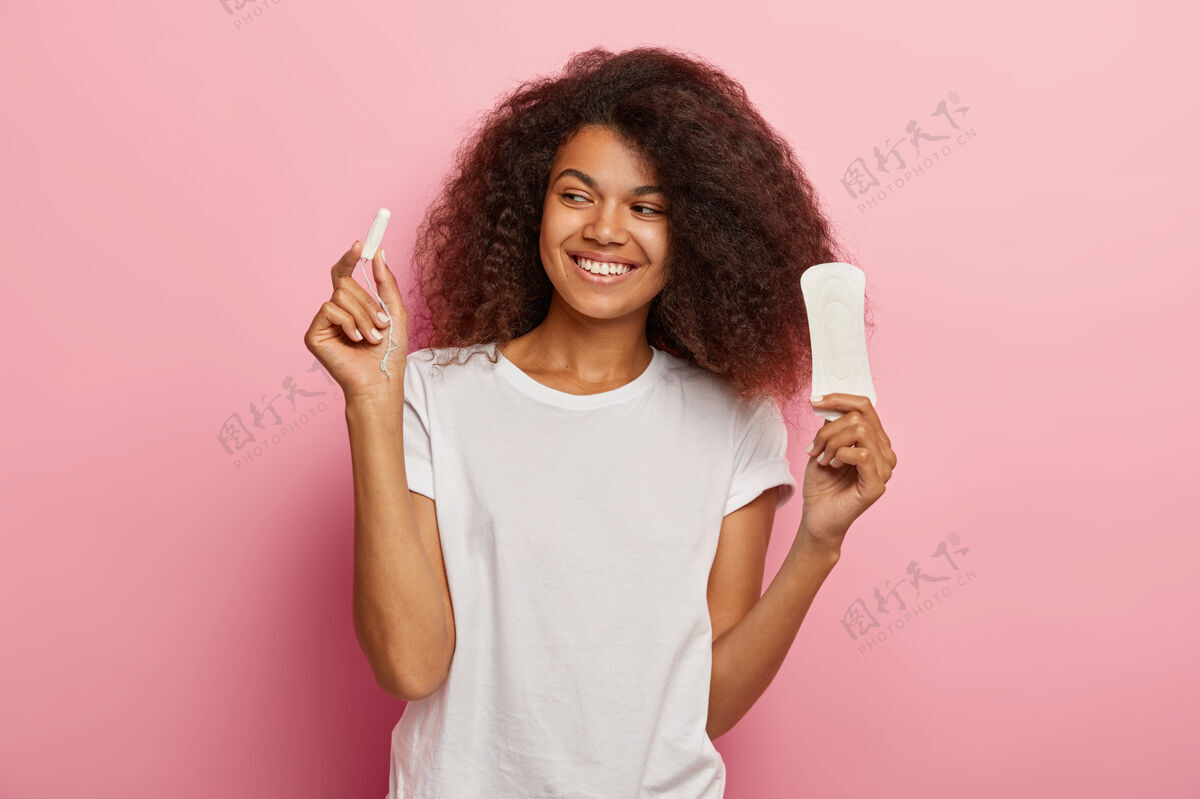 高兴快乐的非洲裔美国妇女拿着卫生棉条和卫生巾 穿着白色t恤 隔离在粉红色的墙上女人 经前综合症室内美式月经