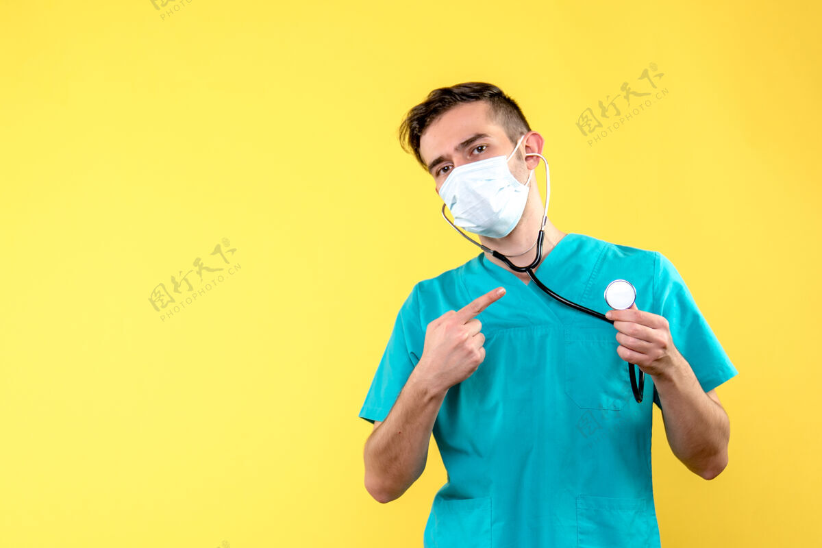防护罩黄色墙上带听诊器和面罩的男医生的正面图健康医院制服