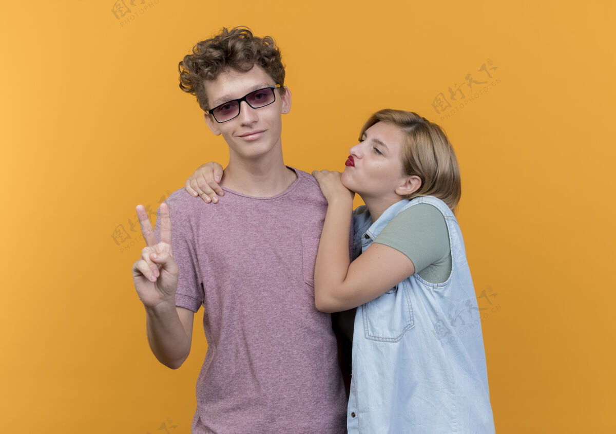 男人一对年轻漂亮的情侣穿着休闲服热恋中的男人和女人站在一起展示着站在橘色墙上的v形标志秀穿站着