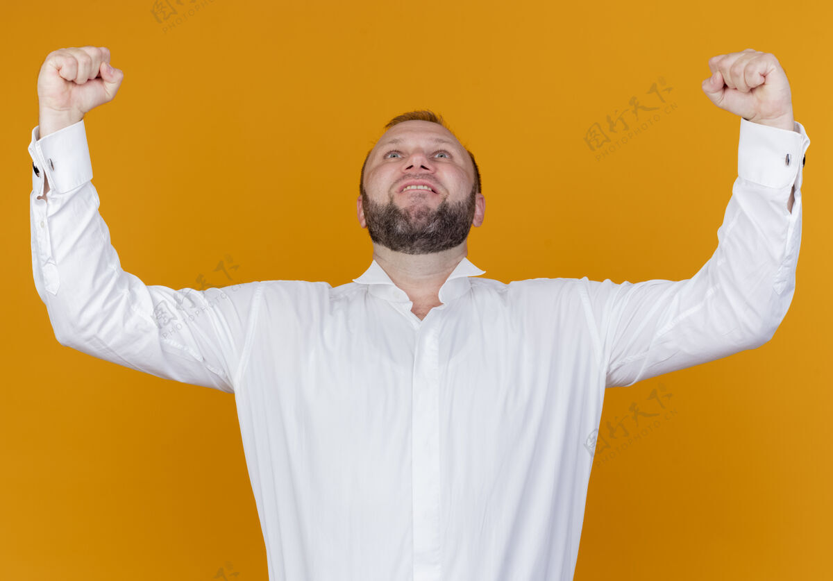 欢喜一个留着胡须 身穿白衬衫的男人举起拳头 站在橙色的墙上为自己的成功而欢呼男人拳头站立