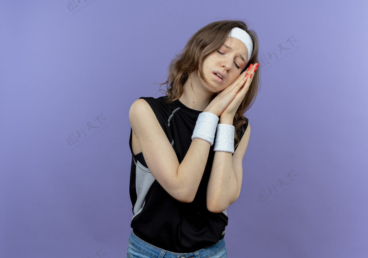 年轻人穿着黑色运动服的年轻健身女孩 头上戴着头巾 双手合十 做着睡觉的姿势 头靠在手心上 想站在蓝色的墙上睡觉保持健康保持健康保持健康