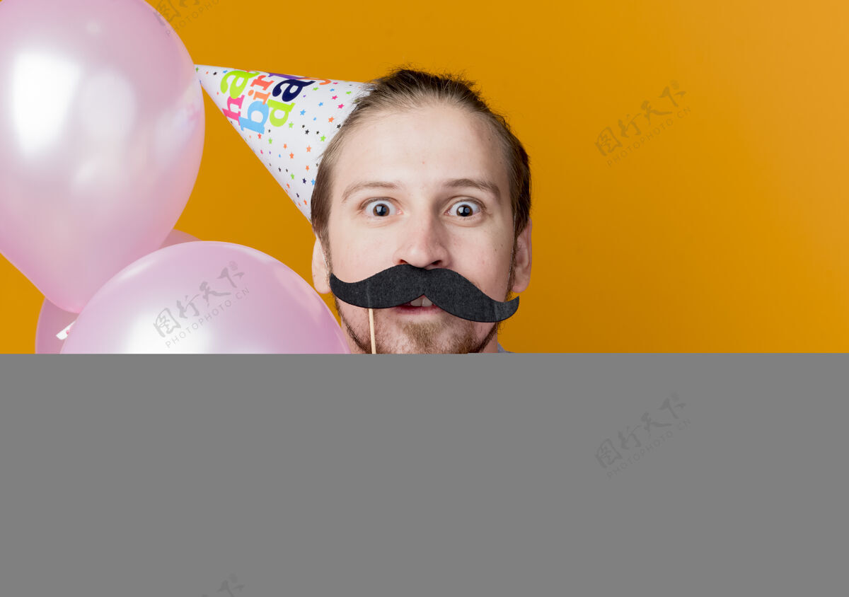 帽子戴节日帽的年轻人手持小胡子派对棒惊讶地站在橙色墙上的气球生日派对概念气球站着男人