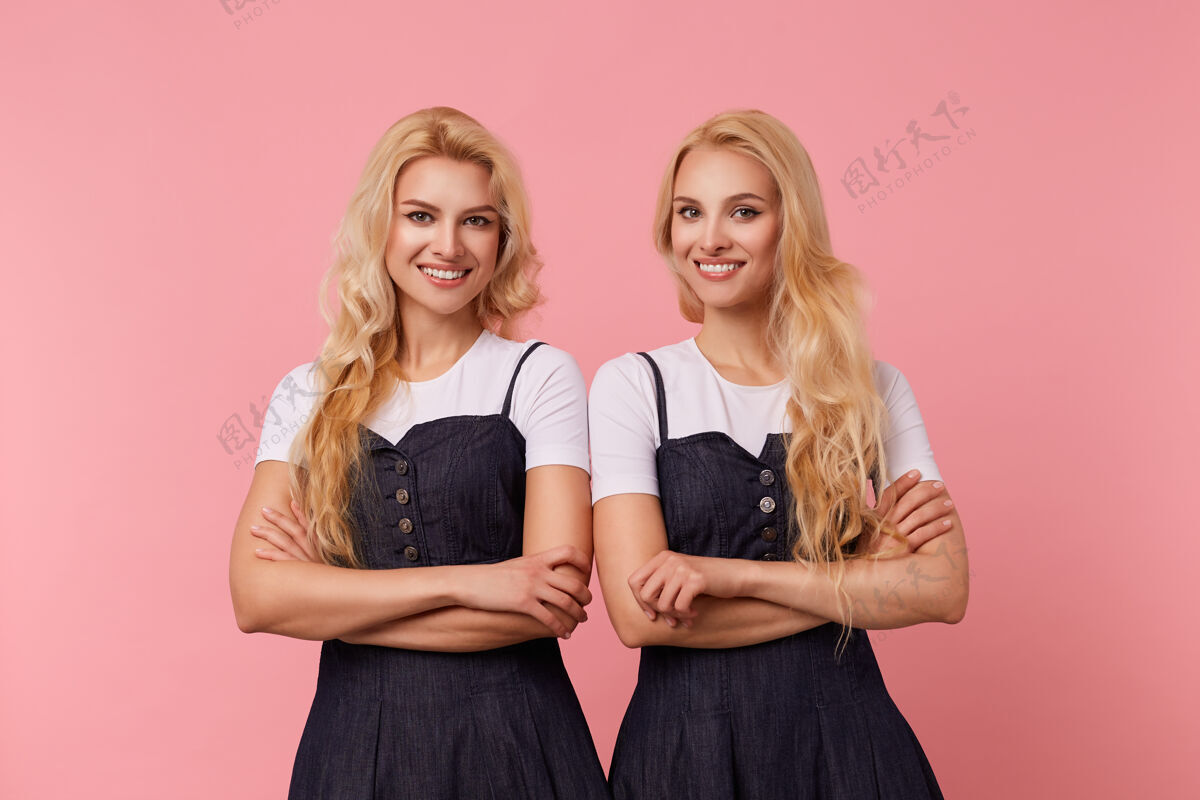 女士一张室内照片 年轻欢快的长发金发女性双手交叉 站在粉色背景上 开心地微笑着看着相机牛仔裤双胞胎女性
