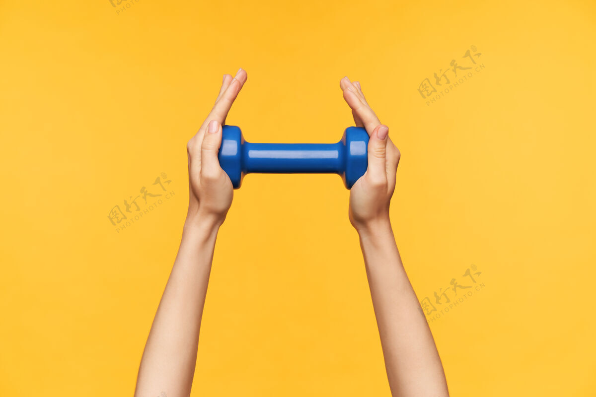 手室内照片 年轻的白皙女士的手被举起 而保持蓝色哑铃在其中 参观健身班和锻炼手臂 隔离在黄色背景皮肤白皙戒指特写
