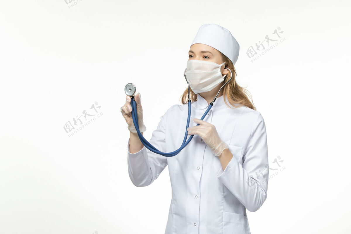 女性正面图年轻女医生穿着医疗服戴防护口罩因冠状病毒在白色表面使用听诊器病毒疾病穿戴