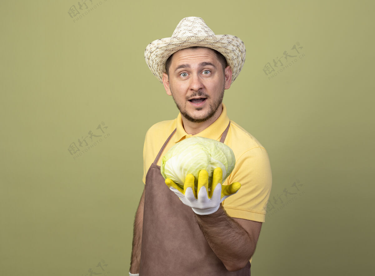 卷心菜年轻的园丁戴着工作手套 穿着连体衣 戴着帽子 在镜头前露出洋白菜的笑脸站着微笑花园