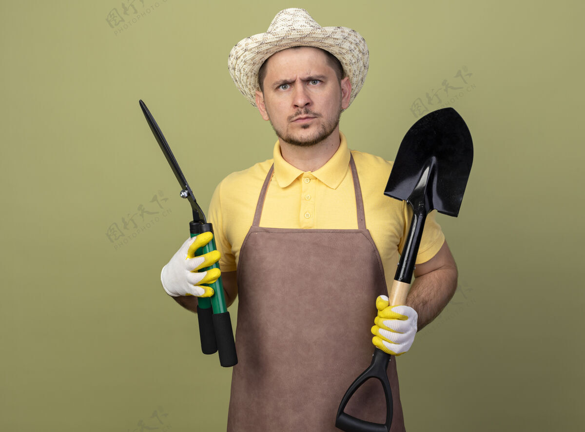 拿着年轻的园丁 穿着连体衣 戴着帽子 戴着工作手套 拿着铲子和树篱剪 表情严肃铲子剪子连身衣