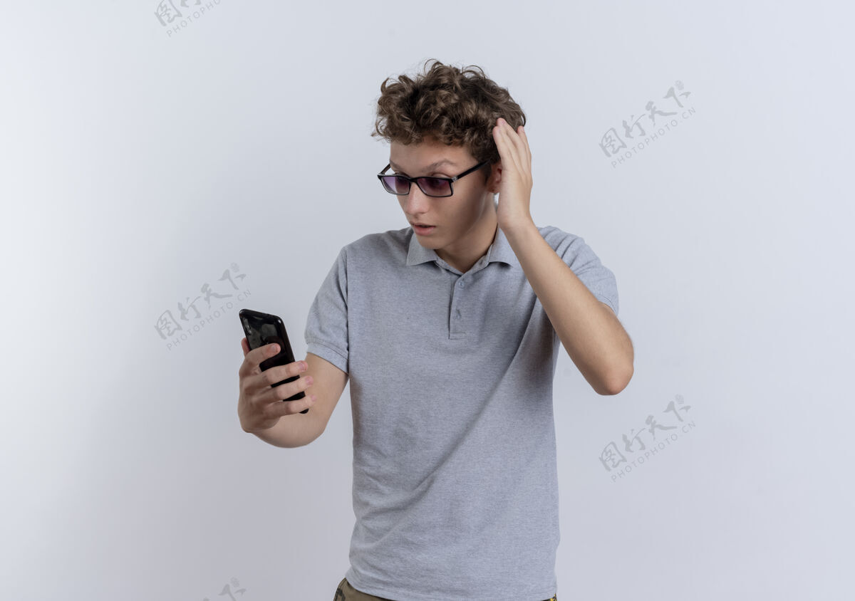 困惑一个穿着灰色马球衫的年轻人站在白色的墙上看着他的智能手机屏幕感到困惑年轻屏幕站