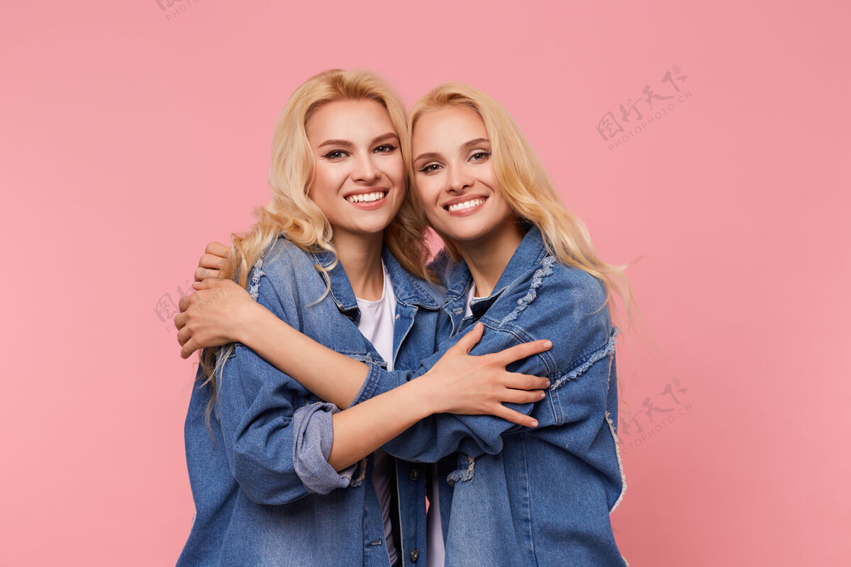 女人快乐的年轻迷人的金发姐妹们 波浪式的发型 温柔地拥抱着对方 愉快地微笑着看着镜头 在粉色背景下与世隔绝表情室内情感