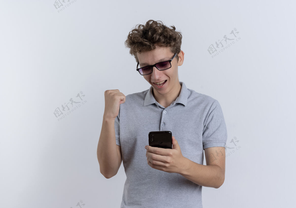 年轻戴着黑眼镜 身穿灰色马球衫的年轻人看着智能手机屏幕 握紧拳头 站在白色的墙上 快乐而兴奋兴奋马球手机