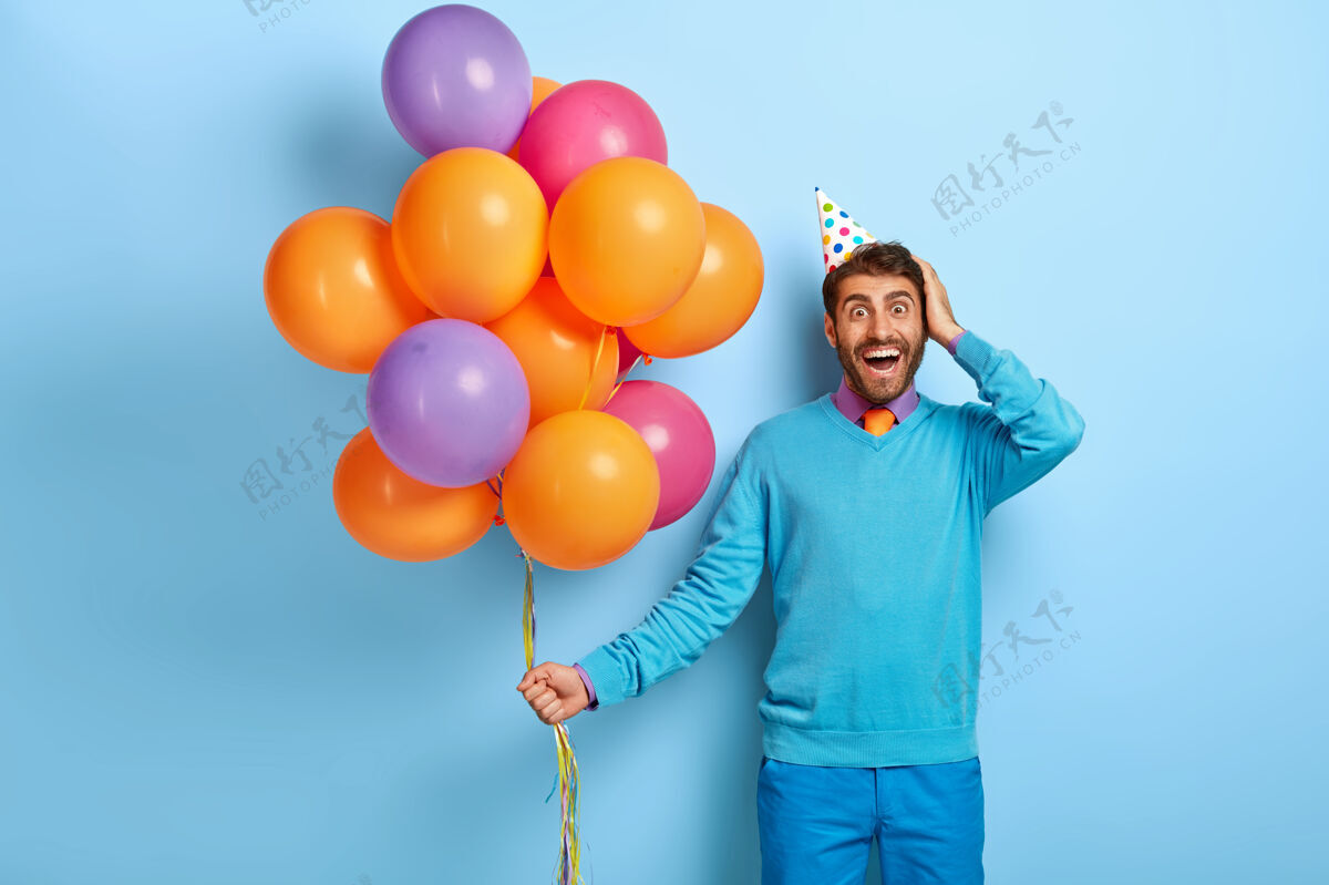 帽子摄影棚拍摄了一个兴奋的家伙戴着生日帽和气球 穿着蓝色毛衣摆姿势姿势满意气球