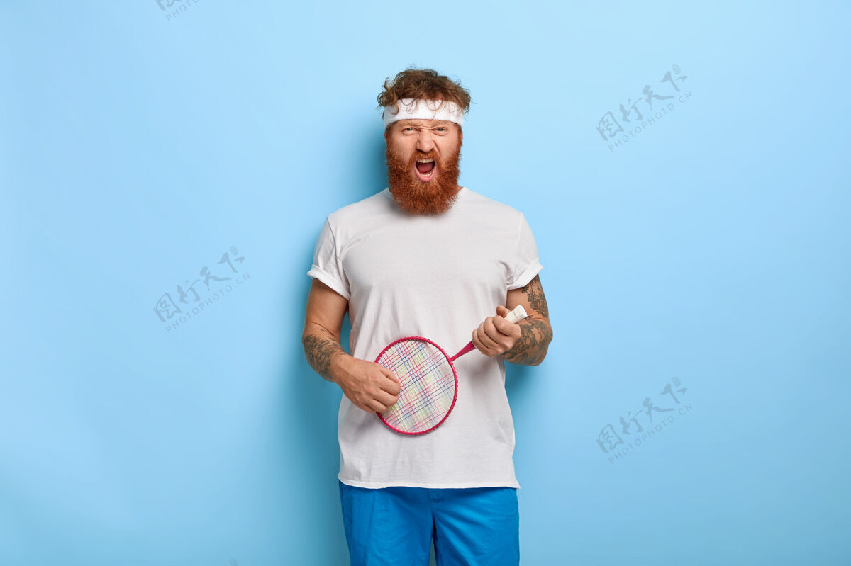 人有趣的红头发网球运动员拿着球拍对着蓝色的墙摆姿势男人网球游戏