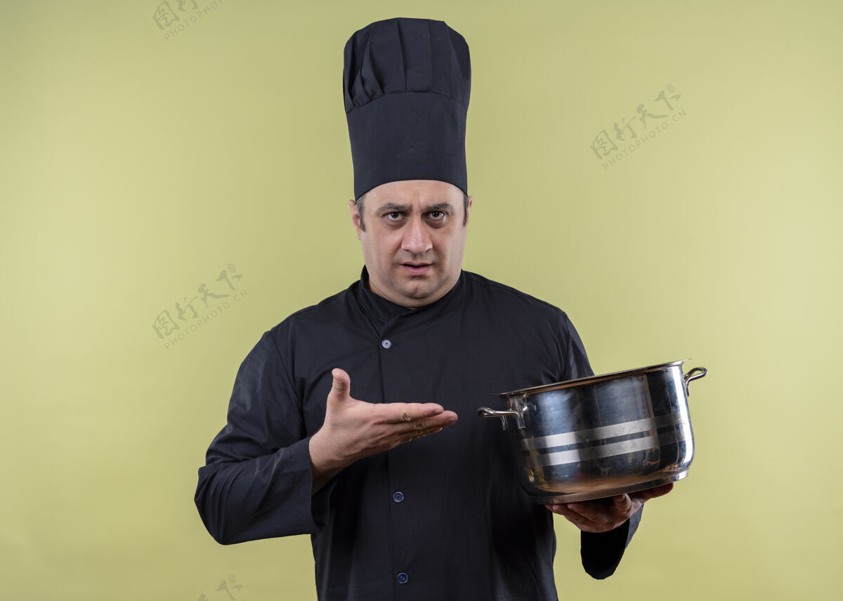 制服男厨师身穿黑色制服 头戴厨师帽 站在绿色背景上 展示着一个胳膊看起来很困惑的平底锅烹饪帽子立场