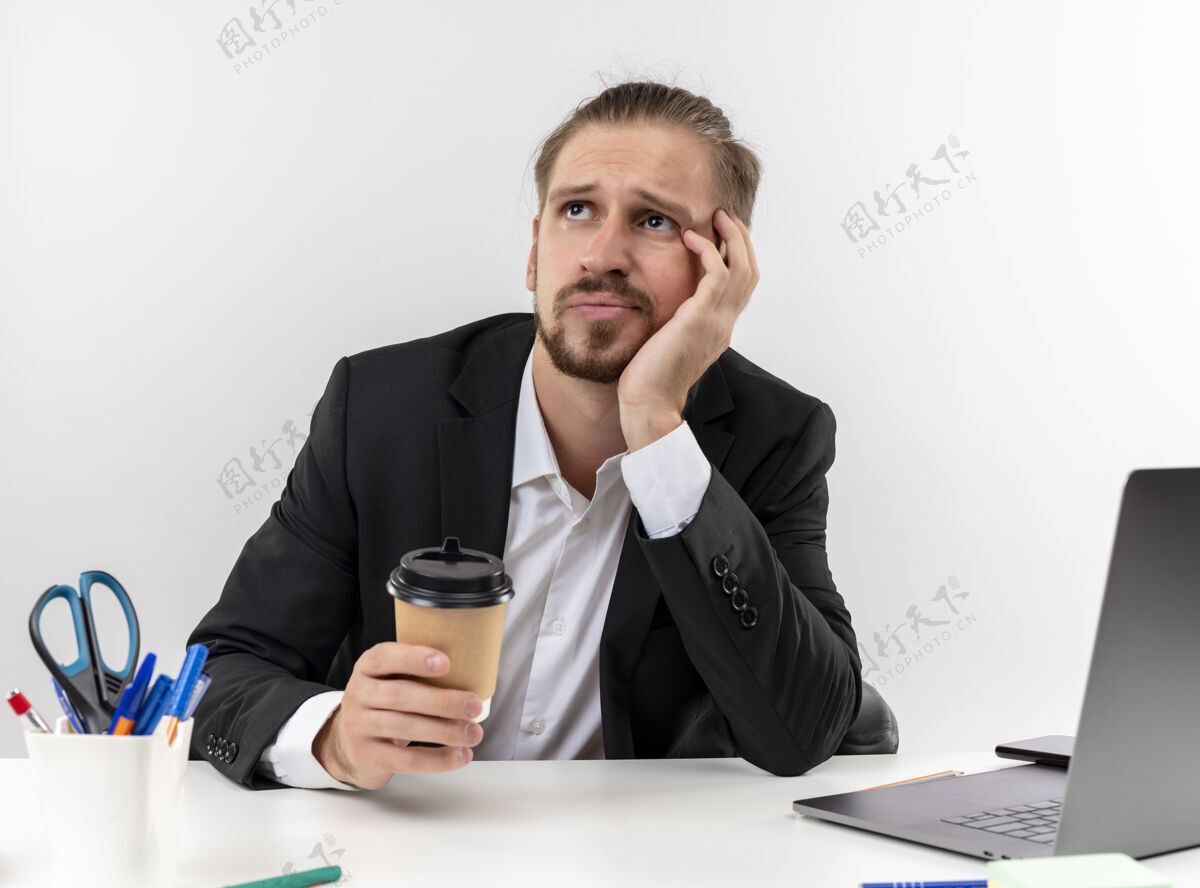 看着穿着西装的帅哥商人拿着咖啡杯在笔记本电脑上工作 看着一边疲惫无聊地坐在白色背景下的办公桌旁工作杯子商务人士