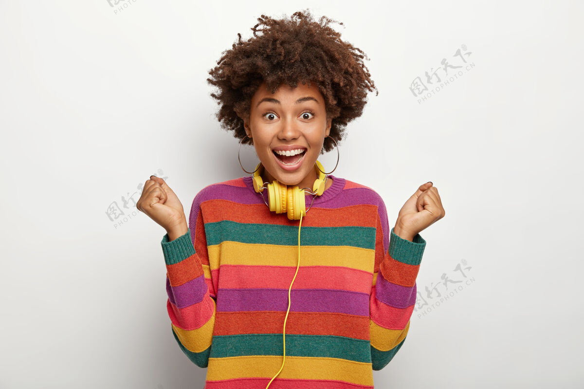 牙关非常高兴的非洲裔美国女士庆祝进球 做出胜利的手势 举起握紧的拳头 有卷发 穿着条纹彩色套头衫 使用耳机连接到一些设备耳机内容黑人