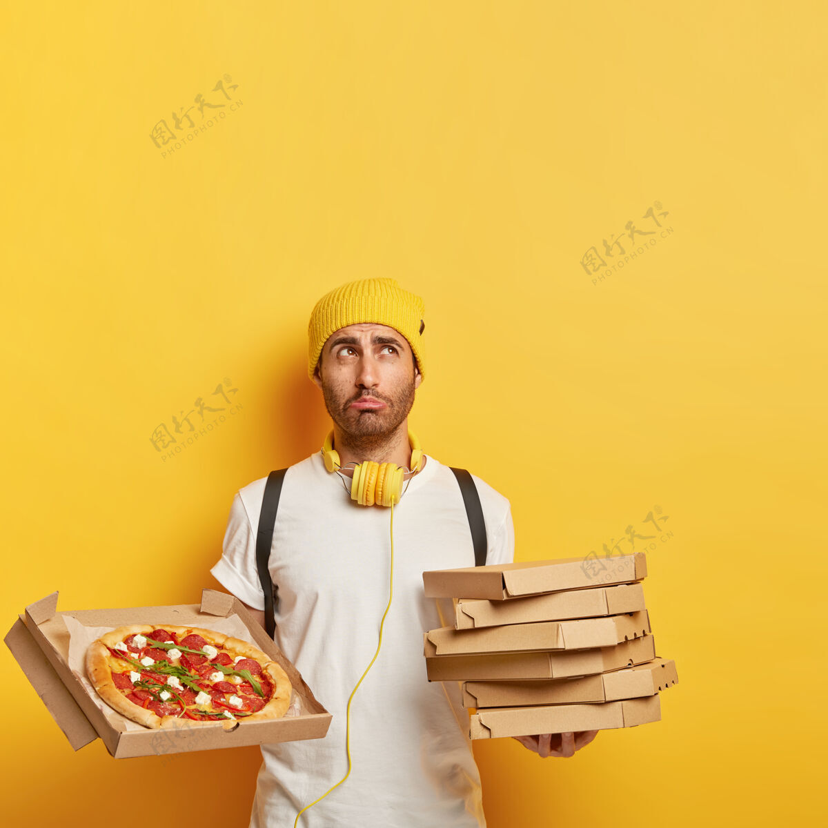 垃圾食品不满意的送货员拿着一堆纸箱 展示美味的奶酪披萨 表情悲伤 戴着黄色帽子和白色t恤站立餐厅纸板