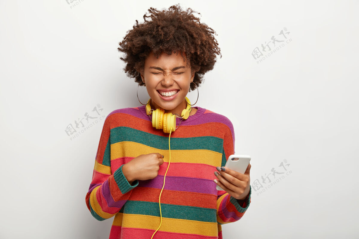 社交喜出望外的快活女人的半身镜头指着自己 拿着手机 表达愉快的情绪 戴着耳环 彩色套头衫 脖子上戴着耳机手机女士应用程序