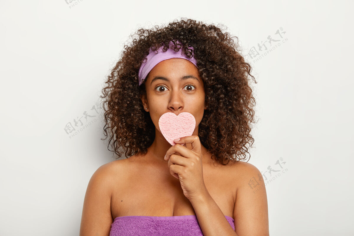 赤裸上身照片中的年轻美籍黑人女子戴着紫色的头巾和毛巾围在身上 用化妆心形海绵捂住嘴 擦脸非洲化妆品年轻化