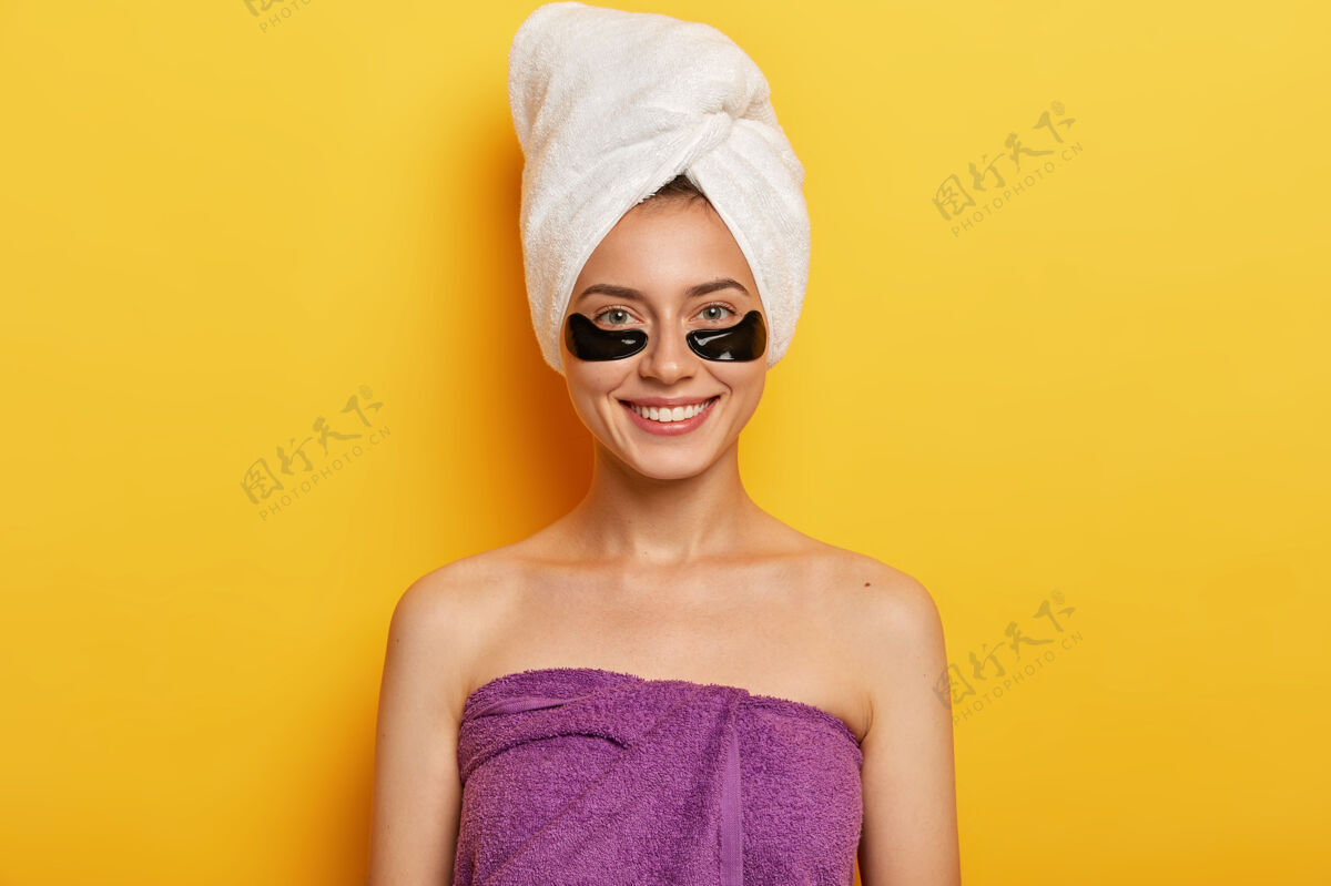 年轻快乐的欧洲女人 面带温和的微笑 有黑色的胶原蛋白斑块 减少黑眼圈的问题 用毛巾包在头上和身上 改善皮肤状况姿势女性面膜