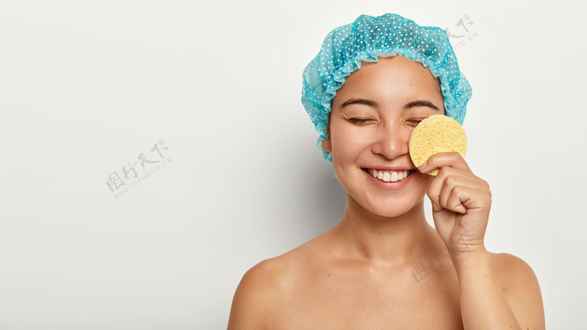 保持快乐的亚洲女人用化妆海绵洗脸 卸妆 闭上眼睛 戴上保护性的淋浴帽健康请特写