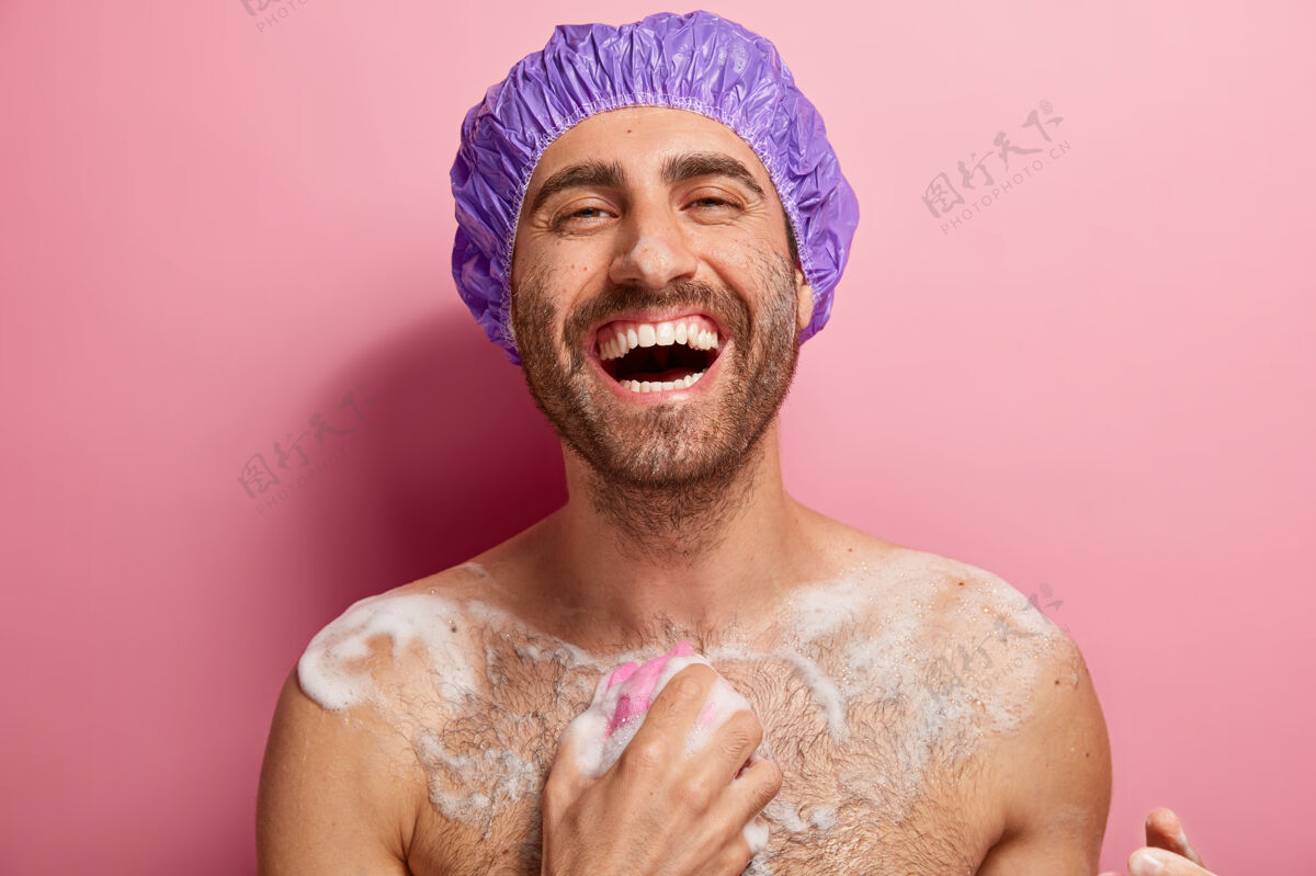 放松欣喜若狂的裸男用凝胶洗脸 身上有肥皂 拿着浴巾 头上戴着浴帽 享受新鲜 洗澡 笑容灿烂 露出洁白的牙齿快乐淋浴阳刚