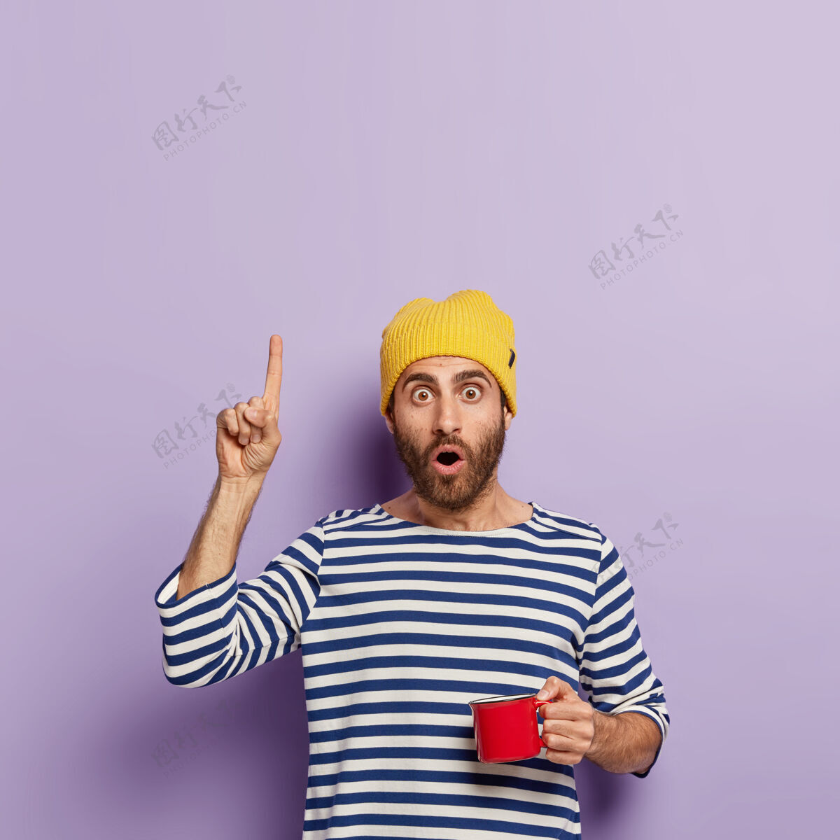 震惊印象深刻的千禧一代用食指向上指 表情震惊 早上喝咖啡 拿着红色的杯子 戴着黄色帽子和条纹水手套头衫 展示了一些东西饮料指向指标