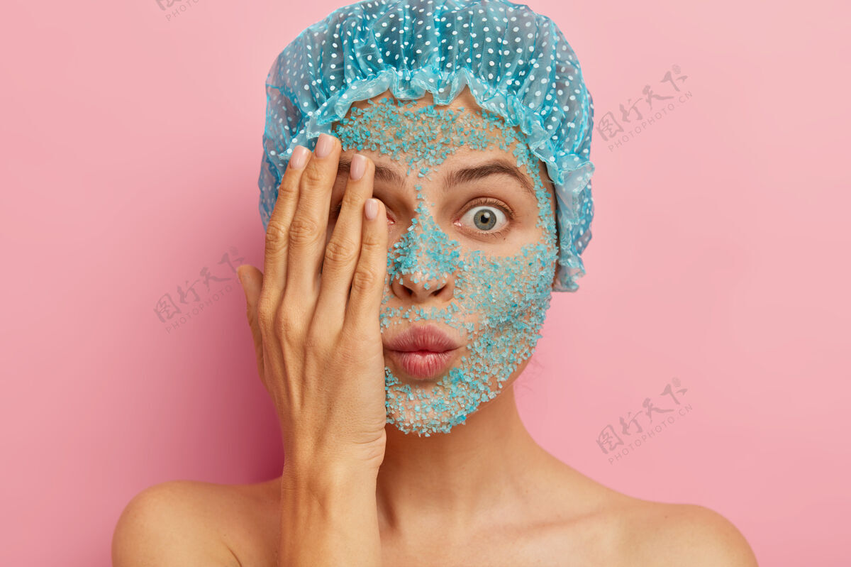 女性脸上涂着蓝色磨砂膏的迷惑不解女子特写镜头 用手捂住一只眼睛 试图隐藏自己 表情呆滞 戴着防护浴帽 想看起来更年轻 赤膊上阵纯洁粉色昏迷