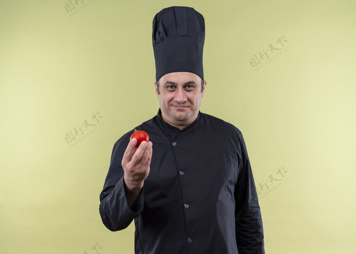 制服男厨师身穿黑色制服 头戴厨师帽 站在绿色背景上看着摄像机 番茄微笑着展示厨师站立